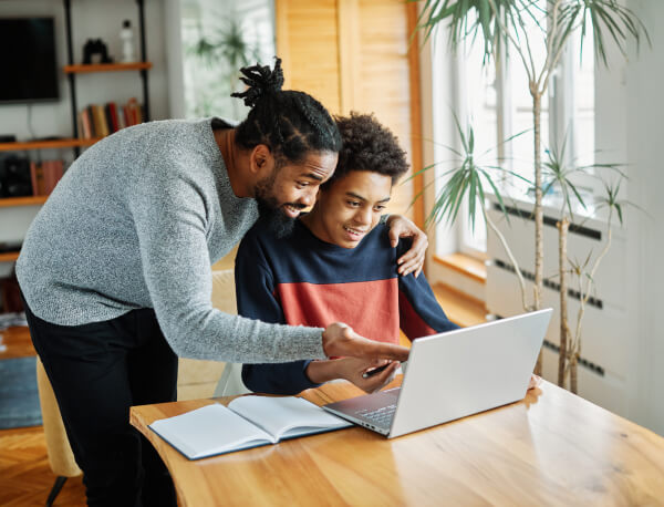 Man Standing Over Son Sitting At Desk Using Computer